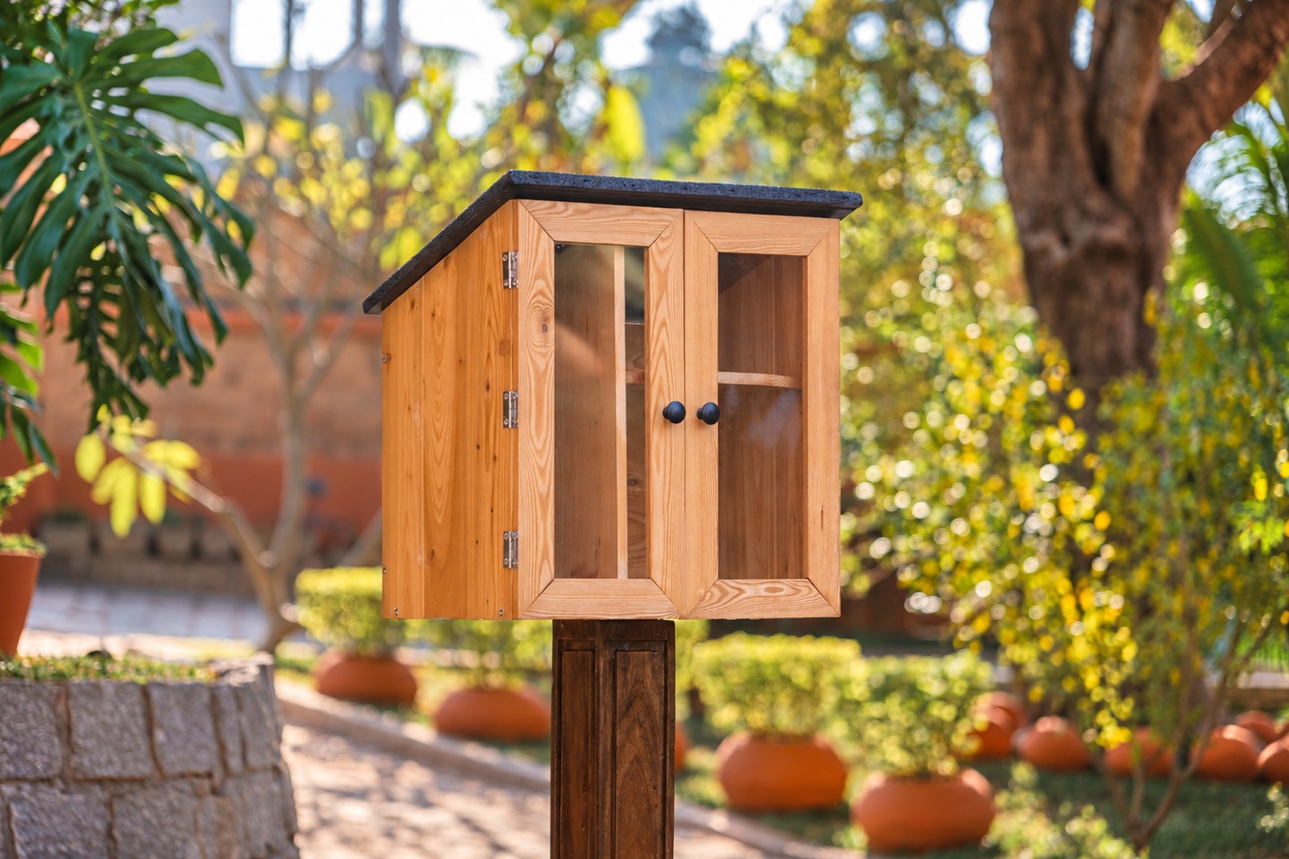 LFL1 wooden outdoor library on a post, with clear doors and a dark roof, in a sunlit garden.